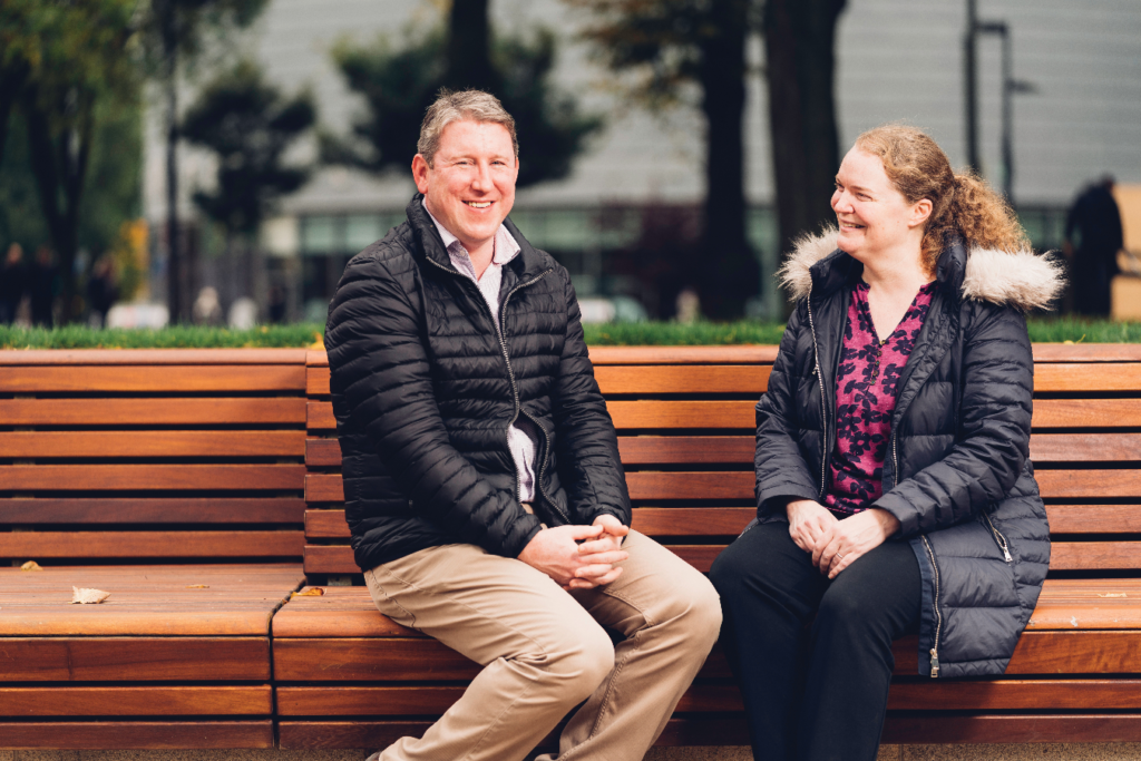 Dr Phil Crosbie, looking at camera smiling and sitting on a bench. Next to him sits Professor Emma Crosbie, smiling and looking at him.
