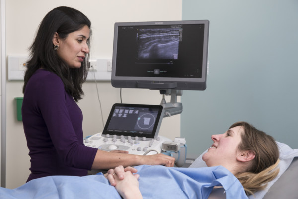 Female sonographer carrying out an ultrasound test on a female patient. Patient is lying in a gown on a bed, while the sonographer presses the ultrasound to her breast and looks at the monitor screen