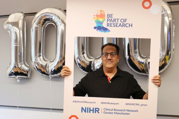 Nazir holding 'Be part of research' sign standing in front of balloons with numbers '1000'