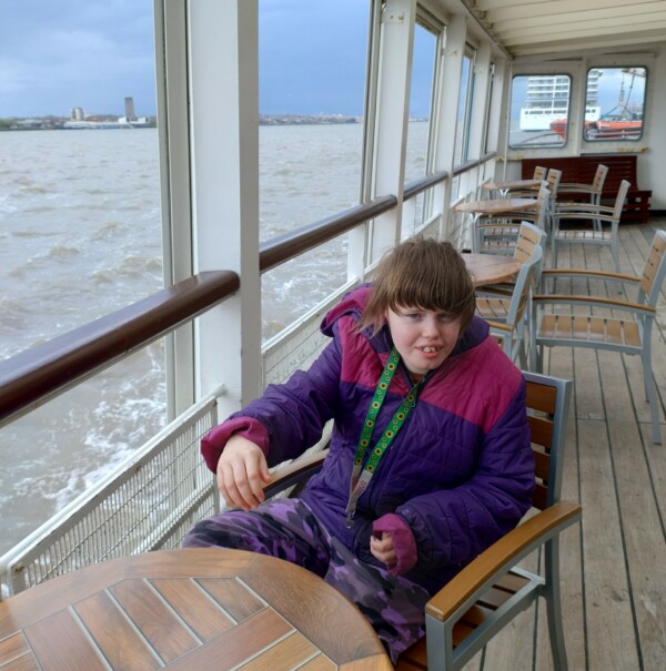 Rose Anderson on a ferry. She is wearing a purple and pink coat and green sunflower lanyard.