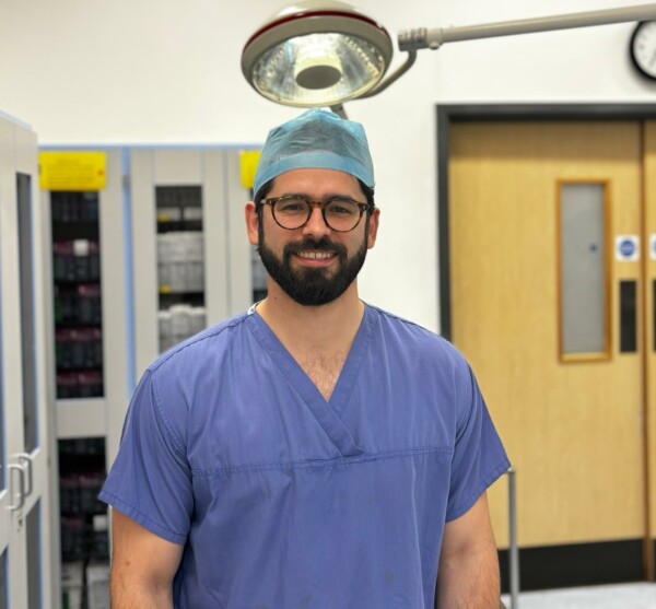 Dr Tom Julian wearing blue scrubs and blue hairnet. He has black round glasses and is standing in a hospital environment